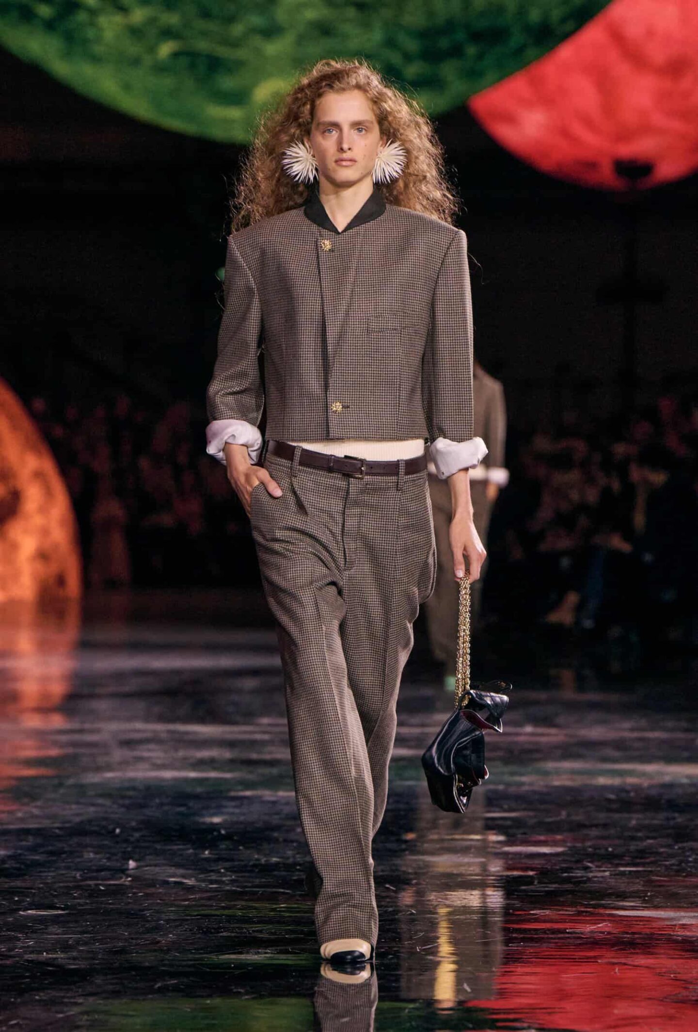 Model wearing a modern Chanel tweed suit in brown tones with a high-collar jacket and tailored pants, styled with statement white earrings, black-and-white slingback heels, and a reinterpreted classic flap bag on the Chanel Fall/Winter 2026 runway.