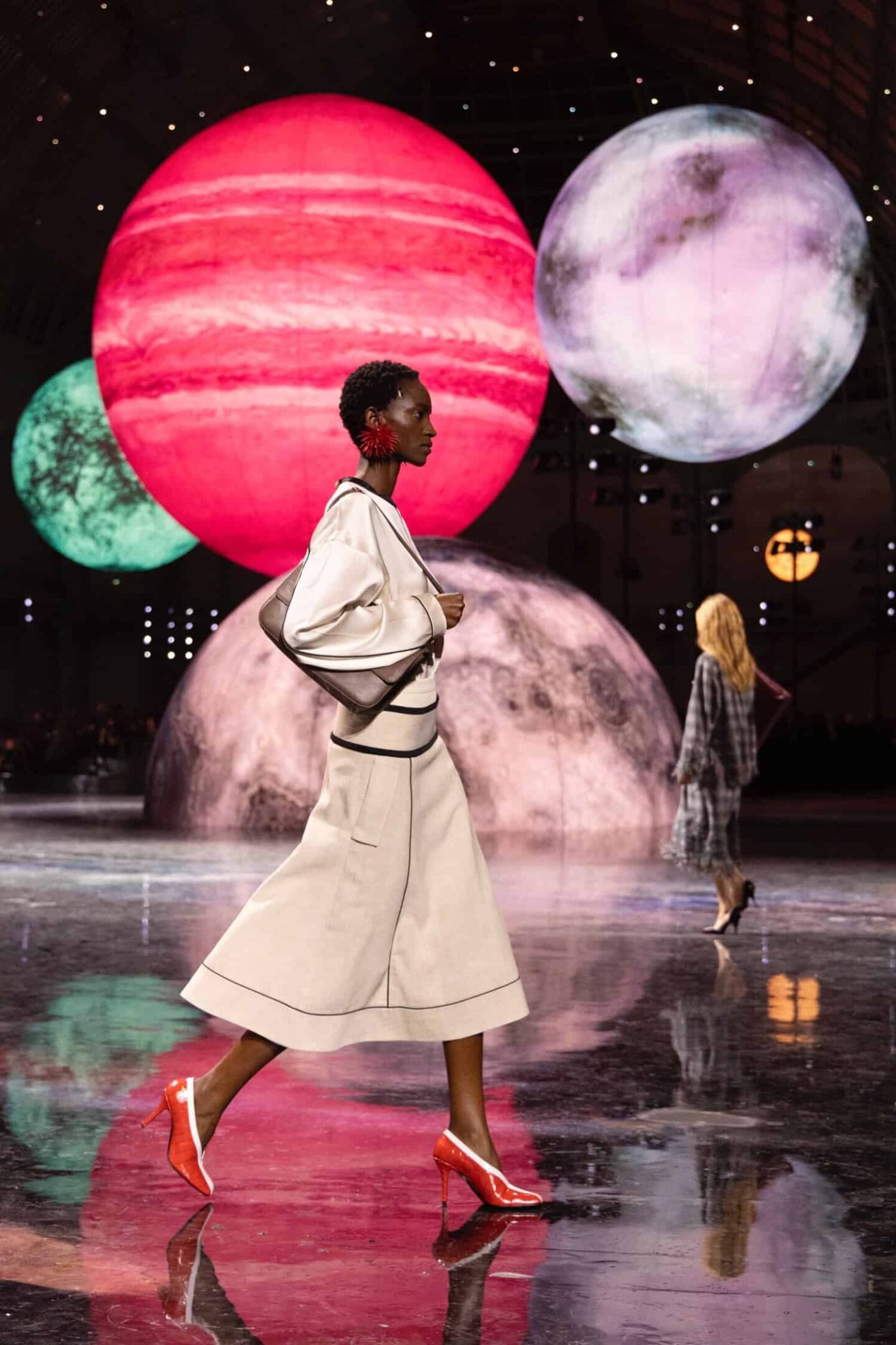 Woman wearing a blouse and beige skirt styled with red croc-embossed square heels with white detailing, a slouchy top-handle shoulder bag, and statement red earrings from Chanel Fall/Winter 2026 runway.
